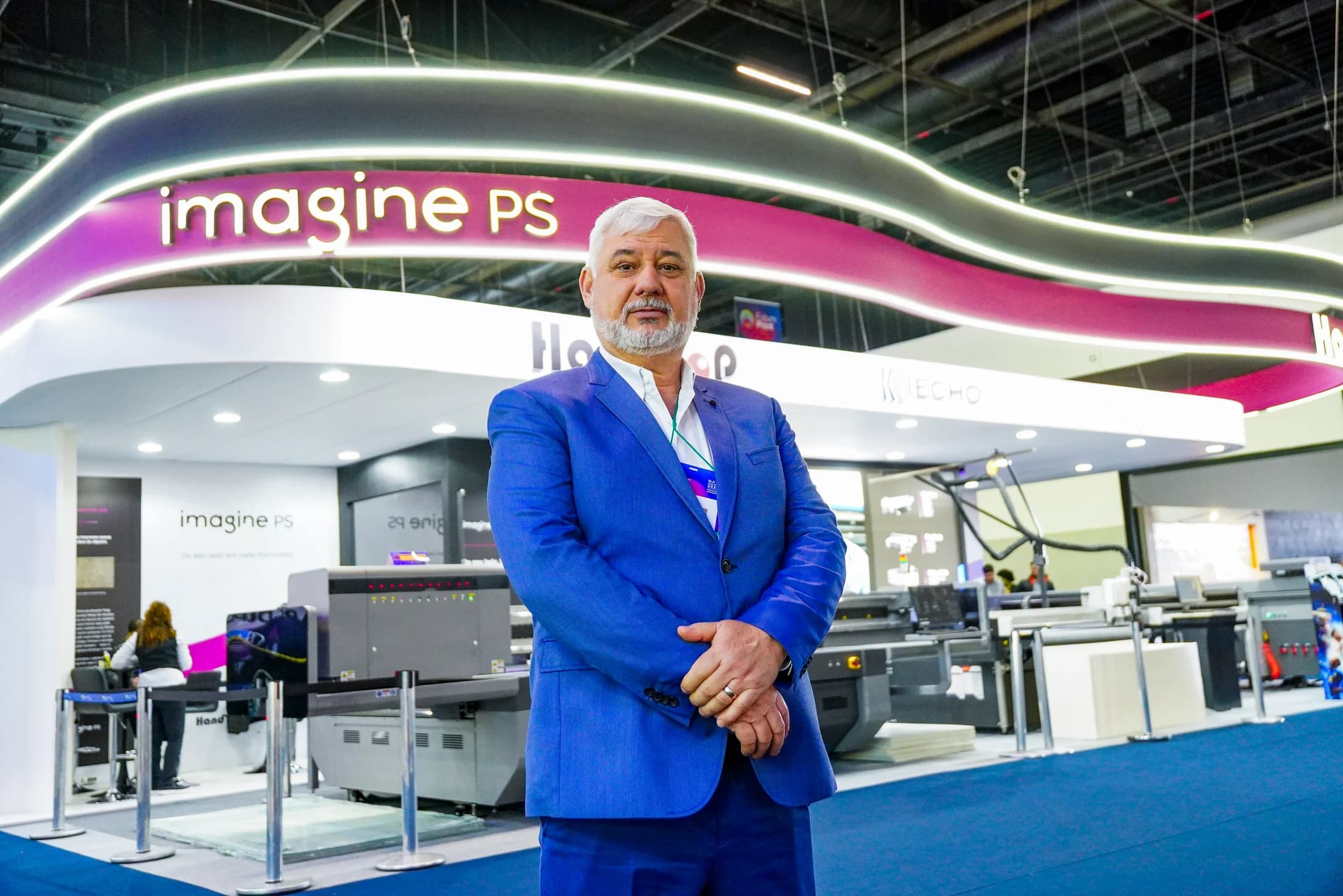 Representante da Imagine PS de terno azul no estande da empresa, com equipamentos ao fundo