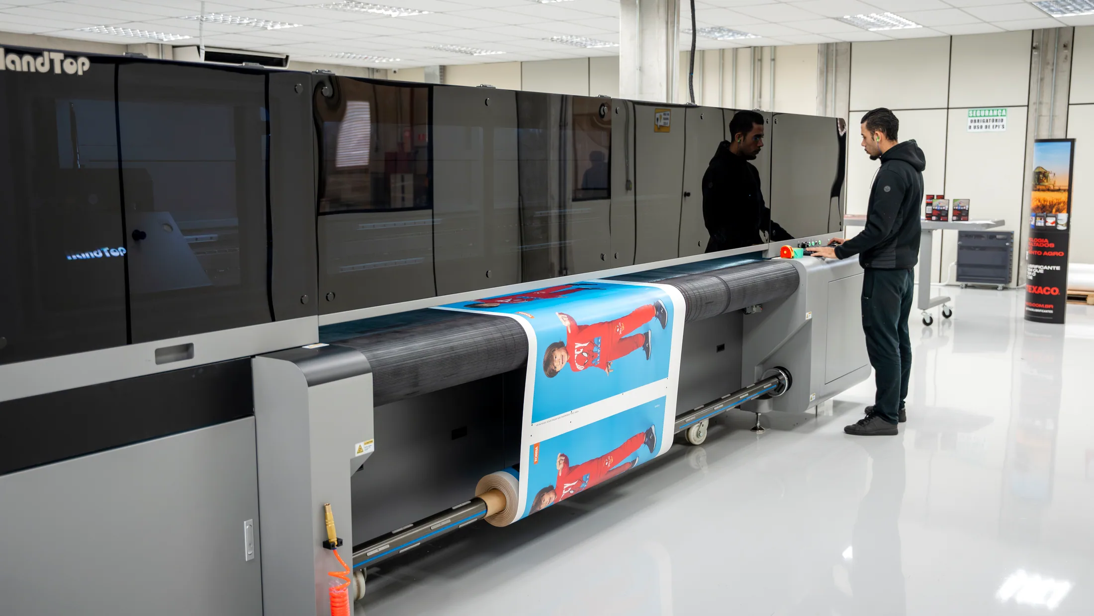 Impressora UV de grande largura produzindo em mídia de rolo, com dois profissionais acompanhando pelo painel