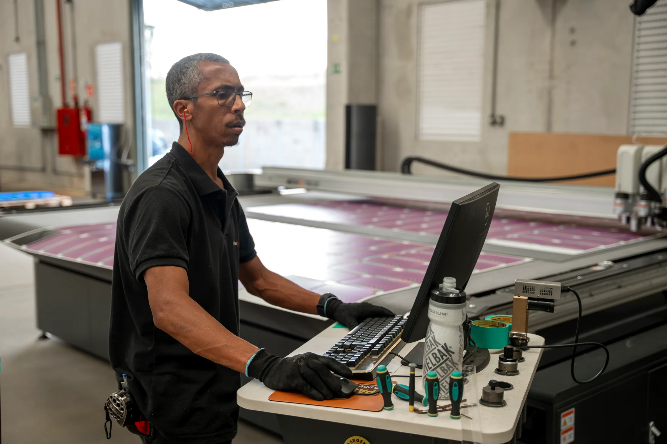 Operador de luvas conduzindo o trabalho pelo computador ao lado de uma mesa de corte digital carregada com material impresso