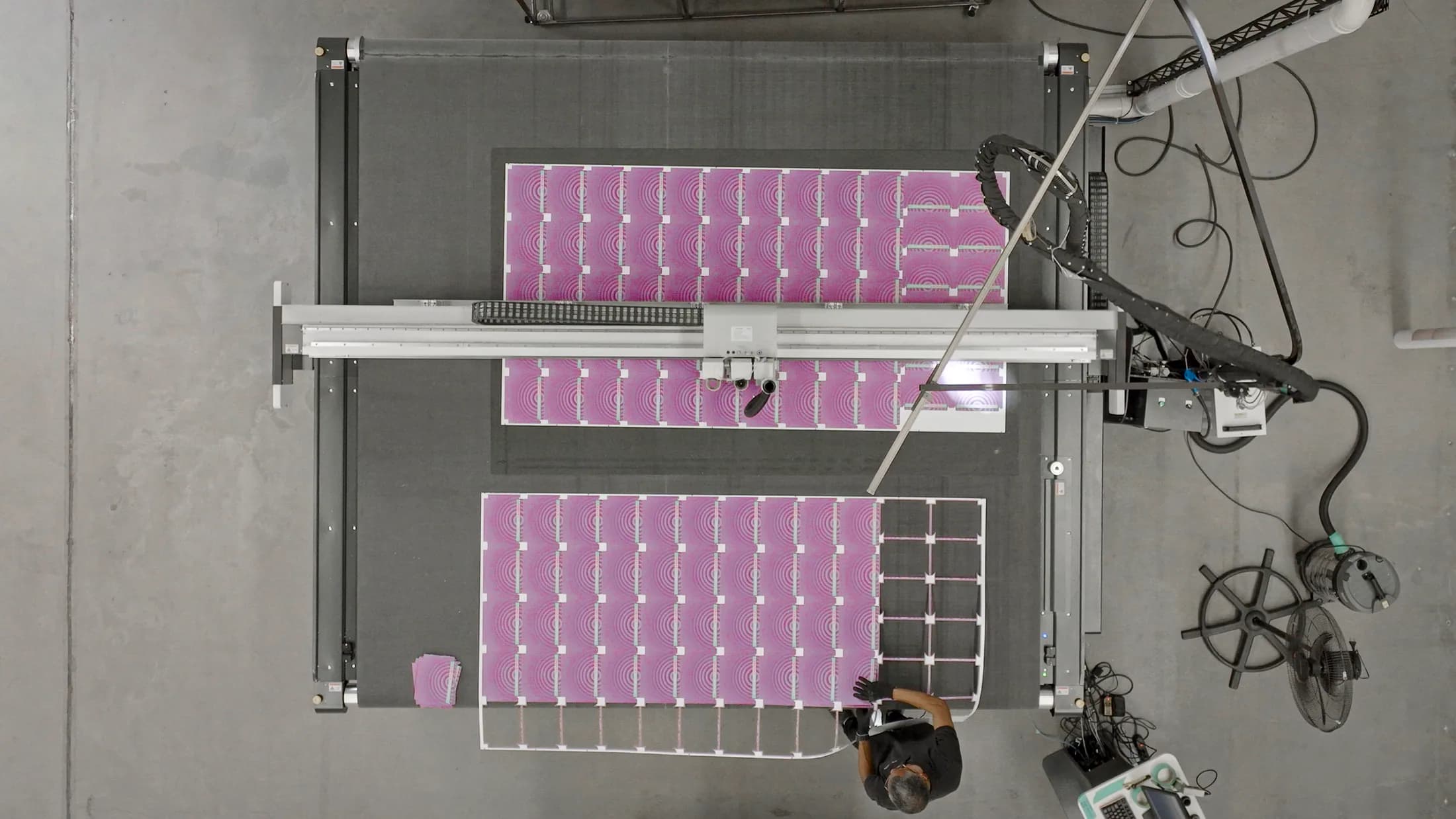 Vista superior de uma mesa de corte digital processando folhas impressas em magenta, com o operador acompanhando ao lado do tapete