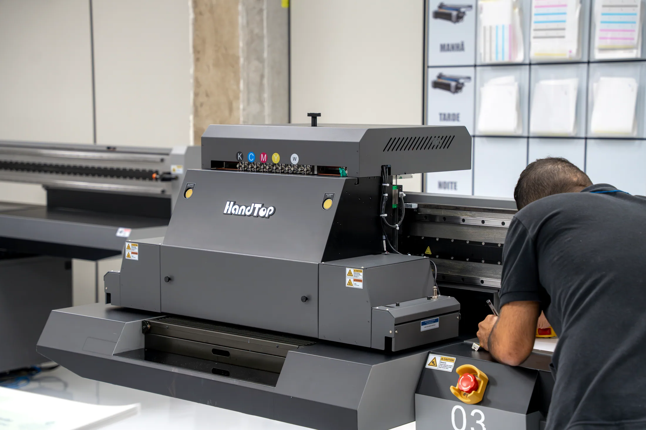 Impressora UV flatbed HandTop em operação, com os canais de tinta identificados no carro de impressão e um operador acompanhando o trabalho ao lado da mesa